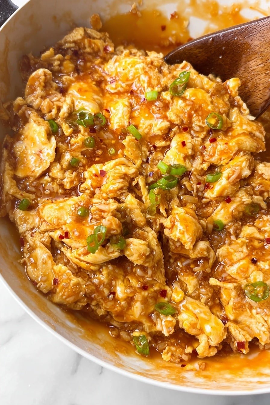 The image shows a close-up of cooked scrambled eggs mixed with small bits of green peppers and onions in a white bowl. The eggs are soft with a light yellow color, slightly soaked in a glossy, orange-brown sauce that covers the mixture evenly. The texture looks creamy with small pieces broken up and coated well by the sauce. The white bowl is partly visible, and a wooden spoon is stirring the food on the top left side. The background is a white marbled surface photo taken with an iphone --ar 2:3 --v 7