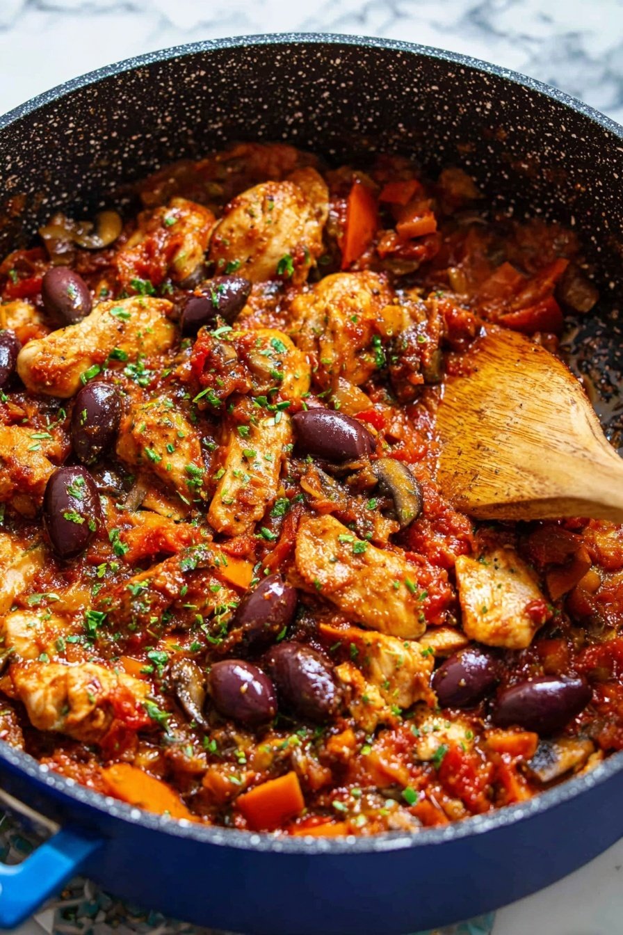 The dish is shown in a deep blue pan with a speckled black interior, filled with cooked chicken pieces in a rich red sauce. There are several layers: the bottom layer is a chunky tomato-based sauce with bits of bell peppers and mushrooms adding splashes of orange, green, and brown. On top are golden-brown cooked chicken pieces scattered throughout, with dark purple olives evenly spread across the dish. Small bits of green herbs are sprinkled over the top, adding freshness. A wooden spoon is stirring inside the pan to the right. The pan rests on a white marbled surface. Photo taken with an iphone --ar 2:3 --v 7