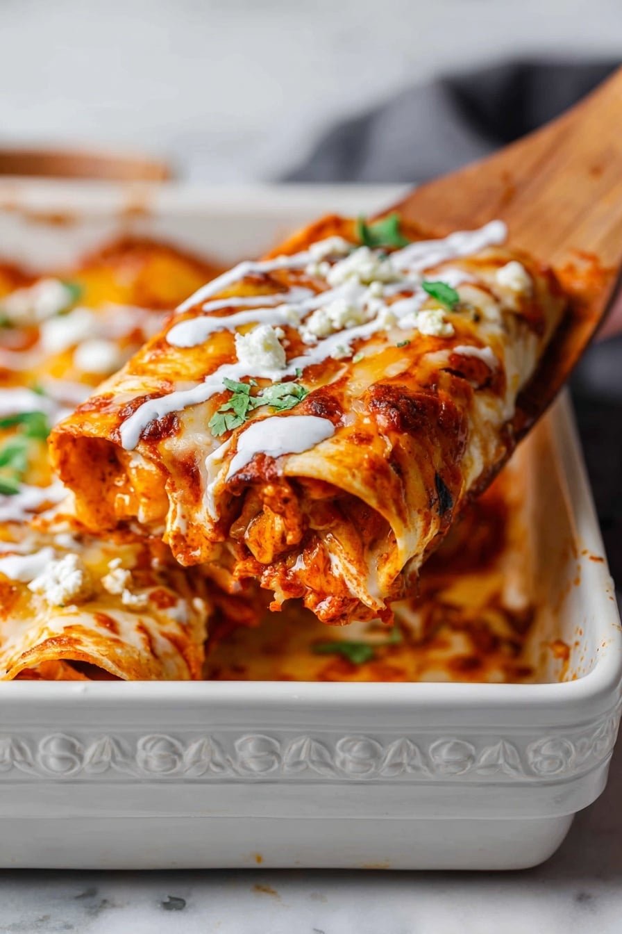 A close-up view of a thick rolled enchilada held by a wooden spatula above a white ornate rectangular baking dish filled with more enchiladas. The enchilada has three visible layers: a soft tortilla as the base, a middle layer of reddish-orange chicken filling mixed with sauce, and a top layer of melted golden-brown cheese with some charred spots. There are streaks of white creamy sauce drizzled across the top, small white crumbles of cheese, and a few green cilantro leaves adding contrast. The dish rests on a white marbled texture surface. Photo taken with an iphone --ar 2:3 --v 7