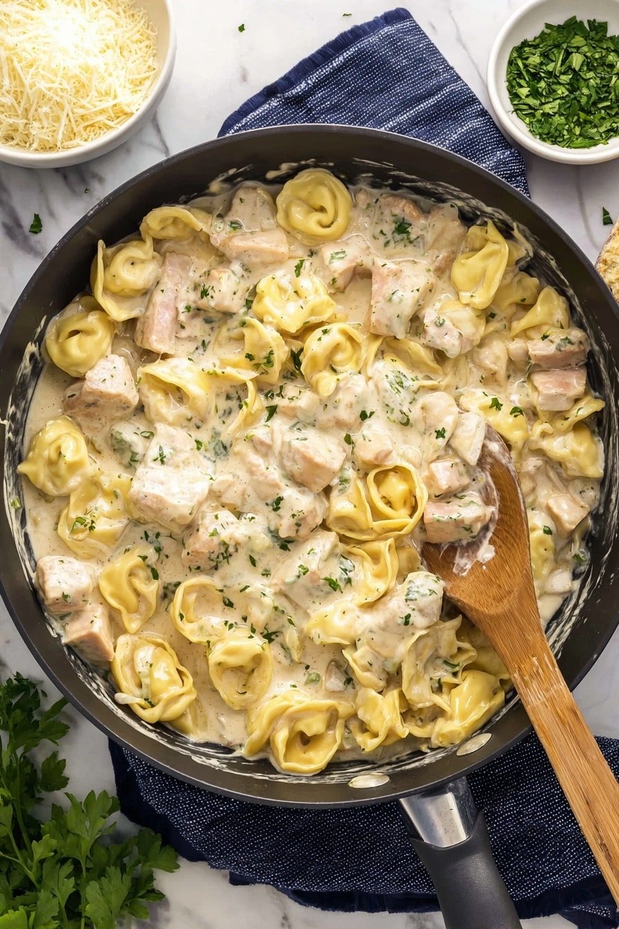 The image shows a large black pan filled with creamy white sauce, small yellow pasta pieces shaped like half moons and circles with ruffled edges, and chunks of pale meat, all mixed together and sprinkled with green herb bits. A wooden spoon rests inside the pan, touching the food. Around the pan are small white bowls, one with chopped green herbs and another partially visible with fine grated cheese. The pan sits on a dark blue and white striped cloth on a white marbled surface. Photo taken with an iphone --ar 2:3 --v 7