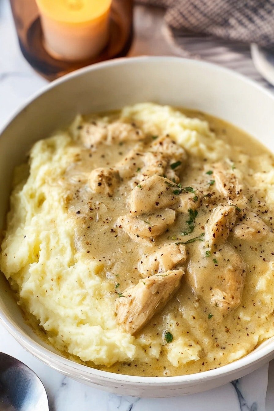 A white plate holds two layers of creamy mashed potatoes, soft and fluffy with a pale yellow color and smooth texture. On top, there is a thick layer of light brown gravy with visible black pepper specks and pieces of tender sliced chicken evenly spread across. The plate sits on a white marbled surface with a lit candle in a glass container blurred in the background. photo taken with an iphone --ar 2:3 --v 7
