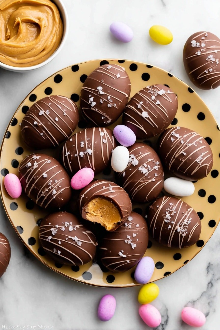 The image shows two chocolate-covered treats stacked on top of each other on a white marbled surface. The top treat has a bite taken out, revealing a pale yellow, crumbly inner filling with a dense texture. The outer chocolate layer is smooth and dark brown with thin curved lines of chocolate drizzled on top. Some small white flakes, possibly sea salt, are sprinkled on the top chocolate layer. Around the treats, there are pastel-colored small candy eggs in soft yellow and light pink shades. In the background, the image is softly blurred with muted light colors. photo taken with an iphone --ar 2:3 --v 7