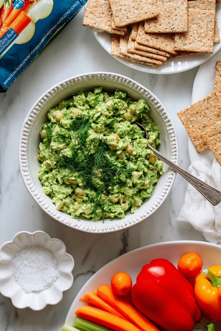 A bowl with a blue pattern contains a bright green chunky dip with visible corn pieces and a sprinkle of fresh green dill on top, with a silver spoon resting inside the bowl. To the left, a white bowl holds light brown, speckled rectangular crackers stacked loosely. Below that, a small white scalloped dish holds coarse white salt. At the bottom left, a white plate shows a mix of fresh bright orange baby carrots, a shiny red bell pepper, and a small orange bell pepper. On the right side, a clear bag labeled