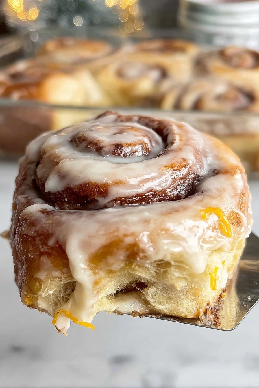 A thick cinnamon roll with three visible layers of soft, light brown dough spiraled tightly with dark brown cinnamon filling. The roll is covered in a creamy, slightly shiny icing that drips slightly at the edges, showing a smooth texture mixed with small bits of orange zest. The cinnamon roll is held by a shiny metal spatula against a white marbled background, with part of the glass pan and other rolls blurred in the back. Photo taken with an iphone --ar 2:3 --v 7