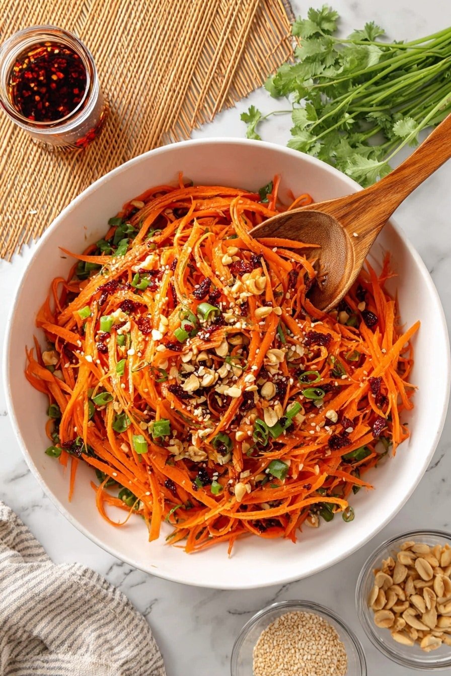 A large white bowl holds a vibrant carrot salad with thin orange carrot strips as the main layer. Mixed in are small green pieces of scallions and cilantro, sprinkled with white sesame seeds and crushed peanuts. Dark red chili flakes and oil are scattered on top, giving a spicy look. Two wooden spoons rest in the bowl, one lifted slightly showing the salad texture. Around the bowl, there is a small glass jar of chili oil on a bamboo mat at the top left, fresh cilantro stems at the top right, and two small clear bowls with peanuts and sesame seeds at the bottom right, all sitting on a white marbled surface. Photo taken with an iphone --ar 2:3 --v 7