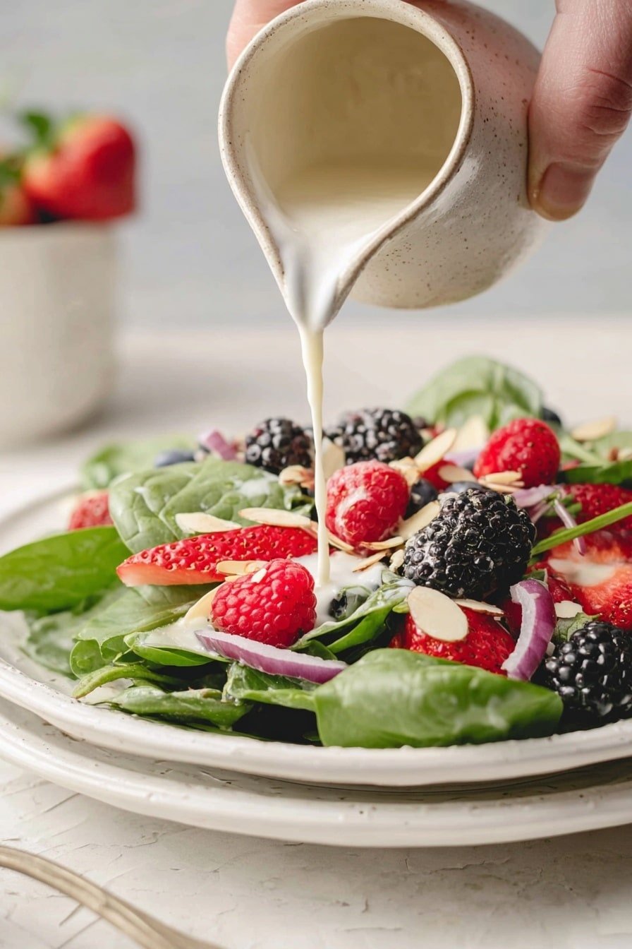 A white plate holds a fresh salad with three main layers: the base layer is bright green spinach leaves spread across the plate, the middle layer consists of red strawberries, black blackberries, and red raspberries scattered on the leaves, and the top layer includes thin purple onion slices and pale almond slices sprinkled evenly. A creamy white dressing is being poured from a textured beige ceramic jug onto the berries, adding a smooth creamy texture to the mix. The scene has a white marbled surface underneath, and in the background, there is a blurred plate with more greens, strawberries, and pink flowers. A woman’s hand is holding the jug. Photo taken with an iphone --ar 2:3 --v 7