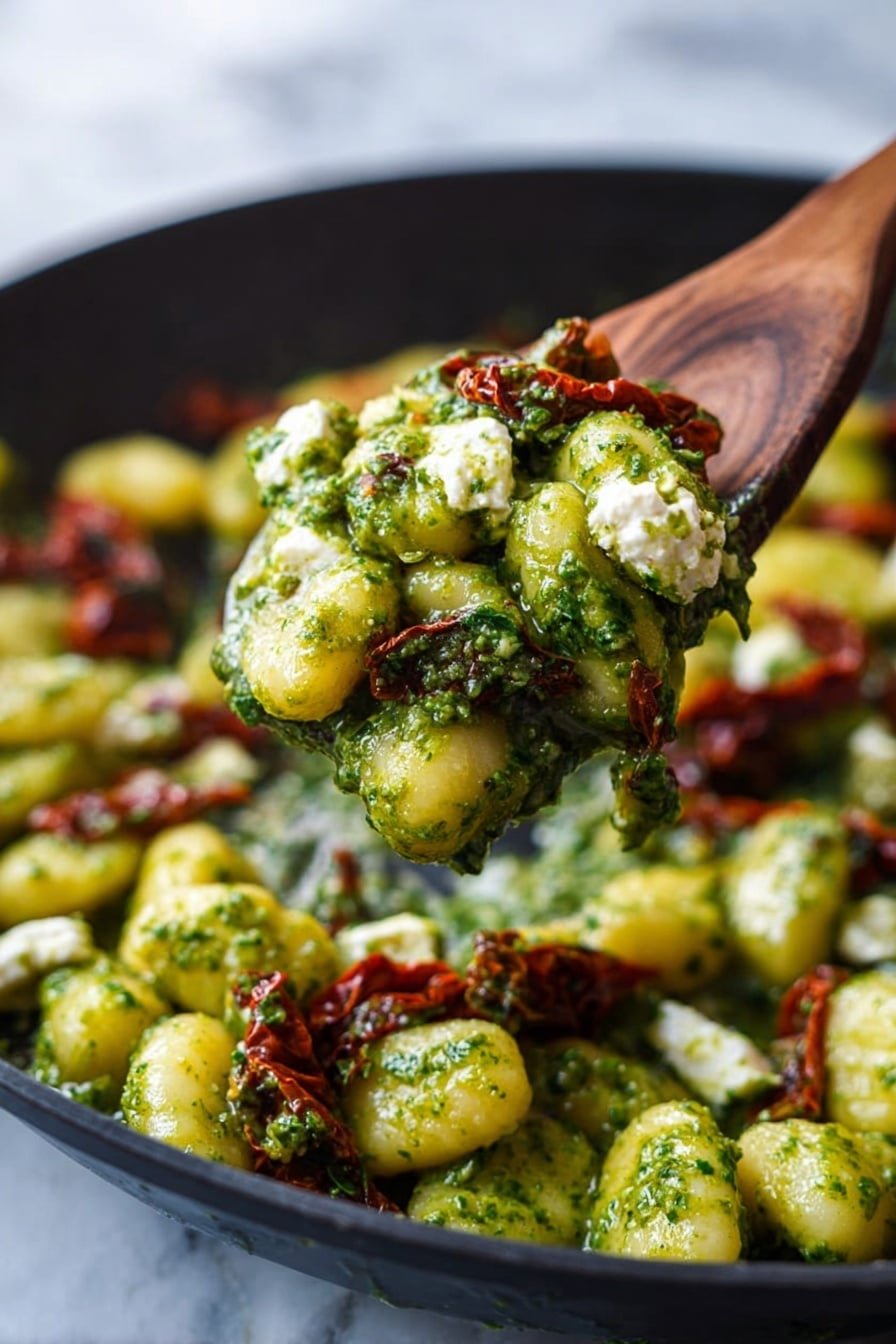 A wooden spoon lifts a spoonful of gnocchi coated in a thick green pesto sauce, showing small white dollops of soft cheese and pieces of sun-dried tomatoes mixed throughout. The gnocchi are light yellow and slightly glossy from the sauce. The background is a black pan filled with more gnocchi, pesto, cheese, and sun-dried tomatoes resting on a white marbled surface. photo taken with an iphone --ar 2:3 --v 7