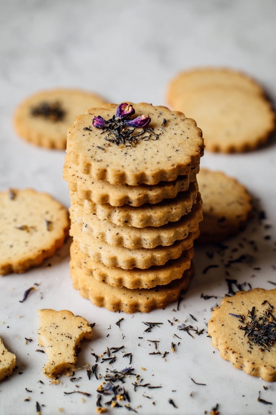 A stack of round, light brown cookies with small dark specks is centered on a white marbled surface, with a few cookies scattered around it. The cookies have a slightly rough texture and scalloped edges, with the top cookie decorated with tiny dried flower petals and small dark crumbs. Some cookies in the background are broken, showing their crumbly inside. Loose small dark tea leaves or crumbs are also spread on the surface around the stack, creating a rustic look. Photo taken with an iphone --ar 2:3 --v 7