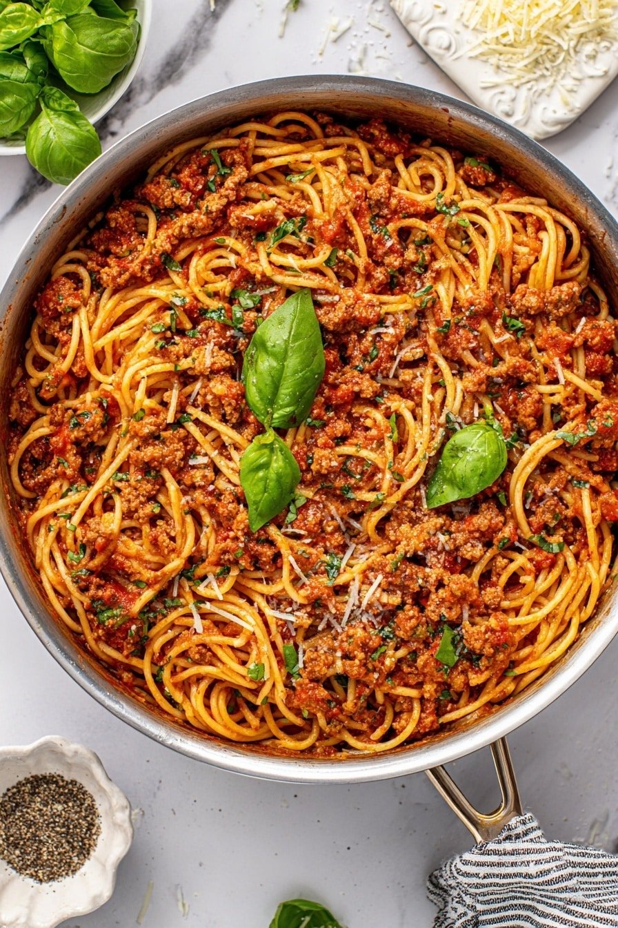 The image shows a large stainless steel pan filled with a layered dish of spaghetti pasta mixed with a rich, chunky tomato sauce containing small bits of cooked ground meat and finely chopped green herbs spread evenly throughout. The sauce is deep red with flecks of green and small white shreds of cheese scattered on top. Bright green basil leaves are placed across the pasta as a fresh garnish. A silver spoon rests in the pan with part of the spaghetti twirled on it. The pan is set on a white marbled surface with some fresh basil leaves in a small ornate white bowl, a small dish of black pepper, and a piece of grated cheese on a white marbled board nearby. Photo taken with an iphone --ar 2:3 --v 7
