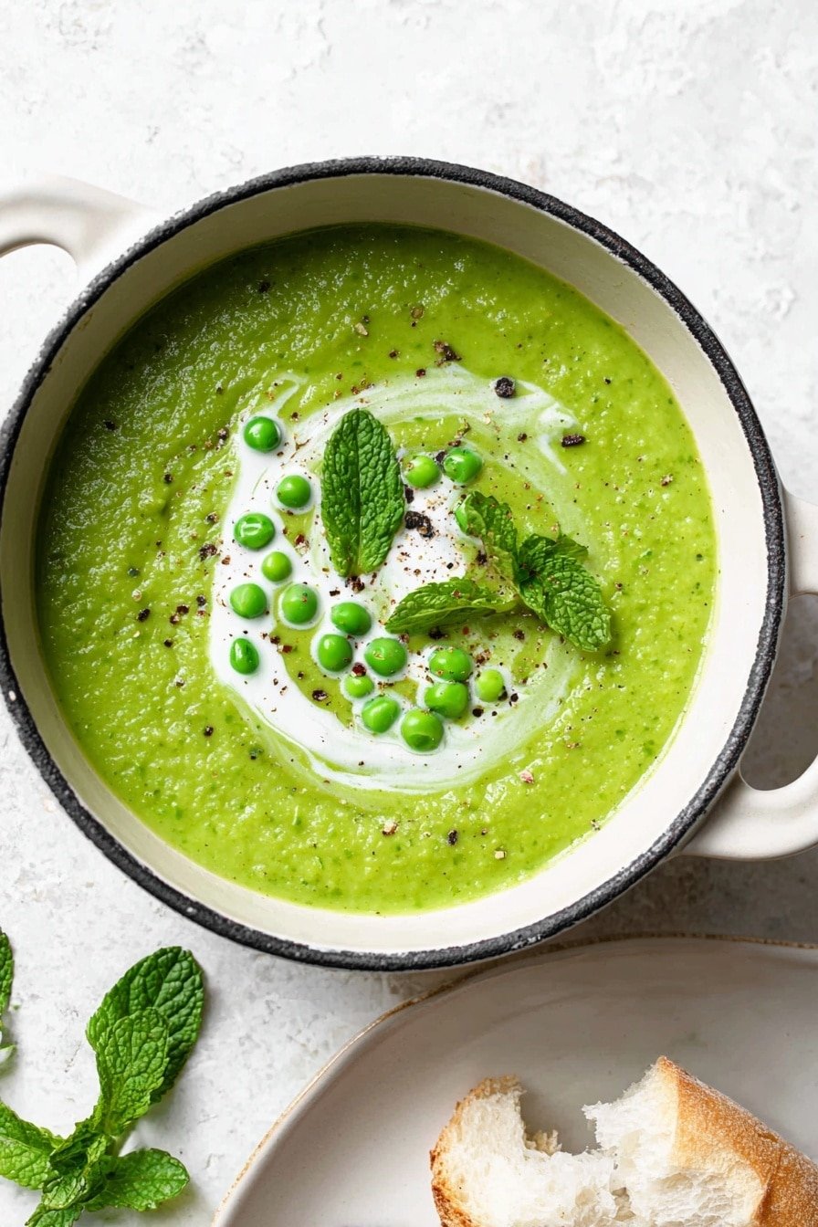 A bowl of thick, green pea soup fills an ornate white bowl, showing a textured surface of blended green peas. On top, there are a few whole bright green peas scattered with a swirl of white cream sauce, and a light sprinkle of black pepper. Near the bowl, fresh green mint leaves rest on a white marbled texture. Part of a white plate with a piece of torn, soft white bread is seen at the bottom right. The photo taken with an iphone --ar 2:3 --v 7