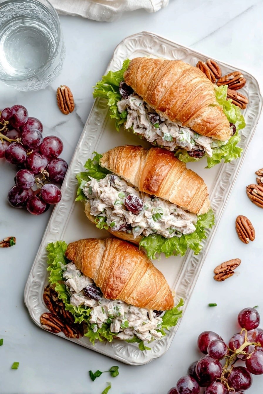 Three croissant sandwiches are placed on an ornate white plate set on a white marbled texture. Each croissant is golden brown and flaky, sliced open to hold a layer of green lettuce at the bottom, followed by a creamy chicken salad mixture with visible chunks of white chicken, dark red grapes, and bits of green celery or herbs. Small clusters of purple grapes and halves of grapes are scattered around the plate and nearby on the white marbled surface, along with pecan nuts and small pieces of green onion. A glass of water is seen in the top left corner. The setting has soft natural light giving a fresh look. Photo taken with an iphone --ar 2:3 --v 7