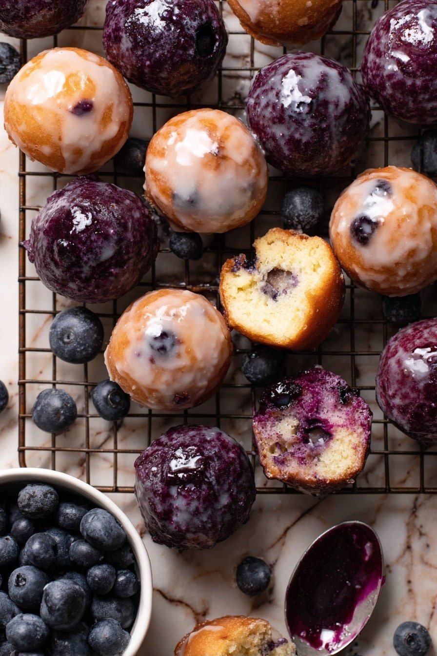 The image shows around twenty small, round mini muffins arranged closely on a metal cooling rack over a white marbled surface. There are two types of mini muffins: one type is light golden brown with a shiny, slightly translucent white glaze and small blueberry pieces visible on top and inside. The other type is dark purple, covered fully in a thicker, wetter dark purple glaze with a few clear drops of white glaze on them, and some blueberries peek out. Two of the mini muffins are cut in half, revealing soft, moist interiors with blueberries inside—one showing mostly golden texture and the other revealing more purple. In the lower left, a white bowl holds fresh blueberries. A silver spoon with dark purple glaze rests on the rack to the right. The lighting is soft and natural, showing the texture of the muffins and glaze clearly. photo taken with an iphone --ar 2:3 --v 7