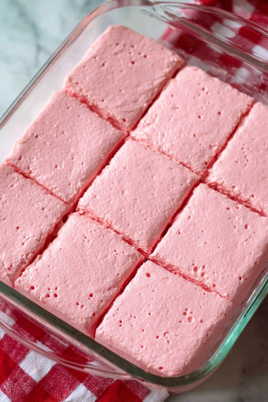 A glass rectangular dish contains a soft, pink creamy dessert evenly spread and divided into nine square portions. The texture looks smooth with small air bubbles throughout, giving it a light and fluffy feel. The background shows a white marbled surface with a red and white checkered cloth partially visible underneath one corner of the dish. The photo taken with an iphone --ar 2:3 --v 7