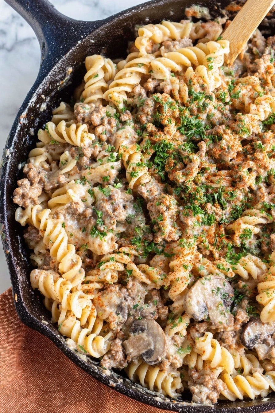 A close-up view of a cast iron skillet filled with a creamy pasta dish made of three main layers: the bottom layer consists of light beige spiral rotini pasta, the middle layer is a mix of small brown crumbled ground meat and sliced mushrooms that are medium brown and slightly glossy, and the top layer is sprinkled with green finely chopped parsley and some red paprika powder for color contrast. The pasta and meat are coated with a light, creamy sauce giving a soft sheen to the whole dish. The skillet rests on a blue cloth placed over a white marbled surface. photo taken with an iphone --ar 2:3 --v 7