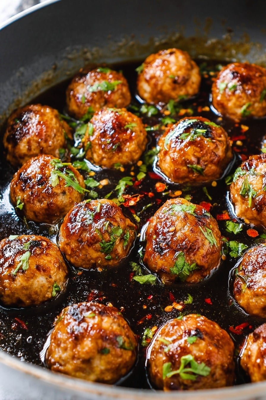 A close-up view of around fifteen shiny round meatballs in a large ornate white pan, covered with a dark brown glossy sauce that pools beneath them. The meatballs are golden-brown with a slightly charred texture and are sprinkled with small green herb leaves and bits of red chili flakes. The pan edges show a slight browning from cooking. The whole scene sits on a white marbled surface. photo taken with an iphone --ar 2:3 --v 7