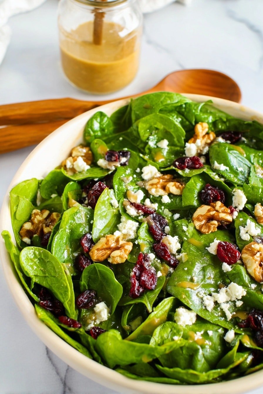 The image shows a wooden bowl filled with fresh green spinach leaves as the base layer, mixed with pieces of light brown walnuts and white crumbled cheese scattered throughout. There are also small dark red dried cranberries mixed in. The bowl is placed on a white marbled texture surface, next to two wooden salad spoons with white handles and some extra dried cranberries scattered around. In the background, there is a blurred small jar with light brown dressing. Photo taken with an iphone --ar 2:3 --v 7