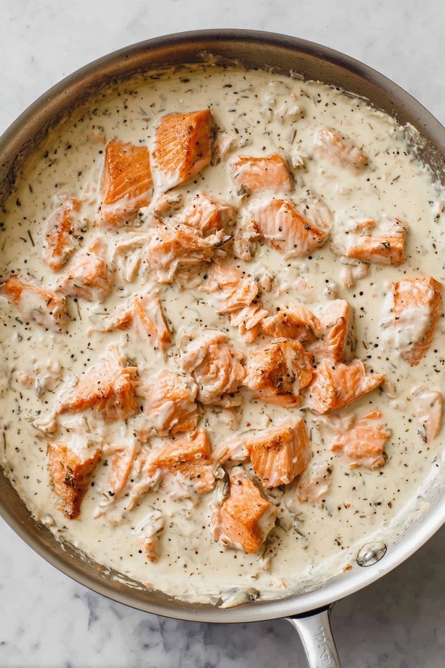 A pan filled with a thick creamy white sauce mixed with small black specks of seasoning, scattered with chunks of cooked orange-pink salmon that have slightly browned edges, creating a contrast with the smooth sauce. The salmon pieces are uneven in size and spread throughout the sauce, which has a light, slightly grainy texture. The pan is placed on a white marbled surface. photo taken with an iphone --ar 2:3 --v 7