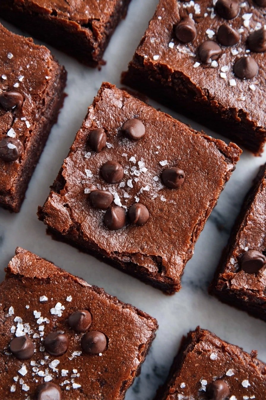 Several thick square brownies are arranged close together on a white marbled surface. Each brownie has one single layer that is dark brown and dense with a slightly textured top. The tops of the brownies are decorated with small dollops of chocolate chips and sprinkled with coarse white salt. The lighting highlights the rich chocolate color and moist texture of the brownies. Photo taken with an iphone --ar 2:3 --v 7