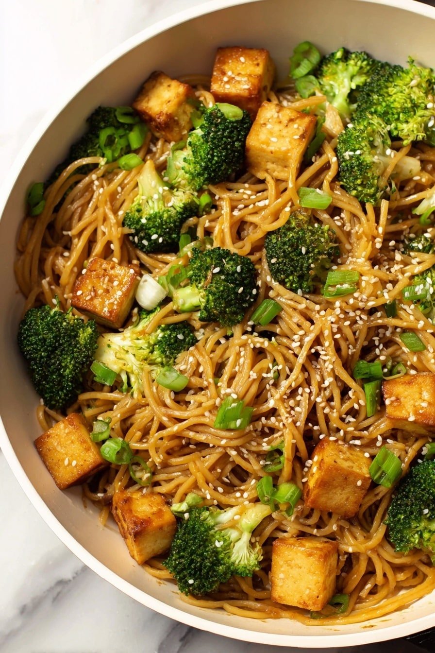 The dish shows a black pan filled with stir-fried noodles as the base layer, light golden brown and glossy with sauce, mixed evenly with bright green broccoli florets and small pieces of green onion scattered throughout. On top are golden-brown tofu cubes, slightly crispy on the edges, and white sesame seeds sprinkled over the whole dish, adding texture contrast. The pan is set on a white marbled surface, with the focus on the colorful mix of noodles, vegetables, and tofu. photo taken with an iphone --ar 2:3 --v 7