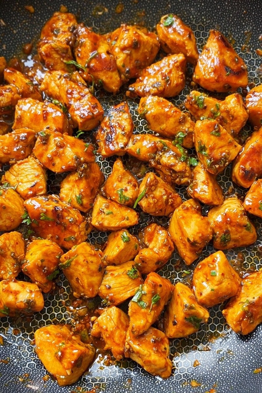 The image shows many small pieces of cooked chicken with a shiny, orange-brown sauce. The chicken pieces are cut unevenly and spread out on the bottom of a metal frying pan with a honeycomb pattern. Some green herbs are sprinkled lightly on top of the chicken pieces. The sauce makes the chicken look juicy and slightly sticky, and the pan's metal texture shines around the edges of the chicken. photo taken with an iphone --ar 2:3 --v 7