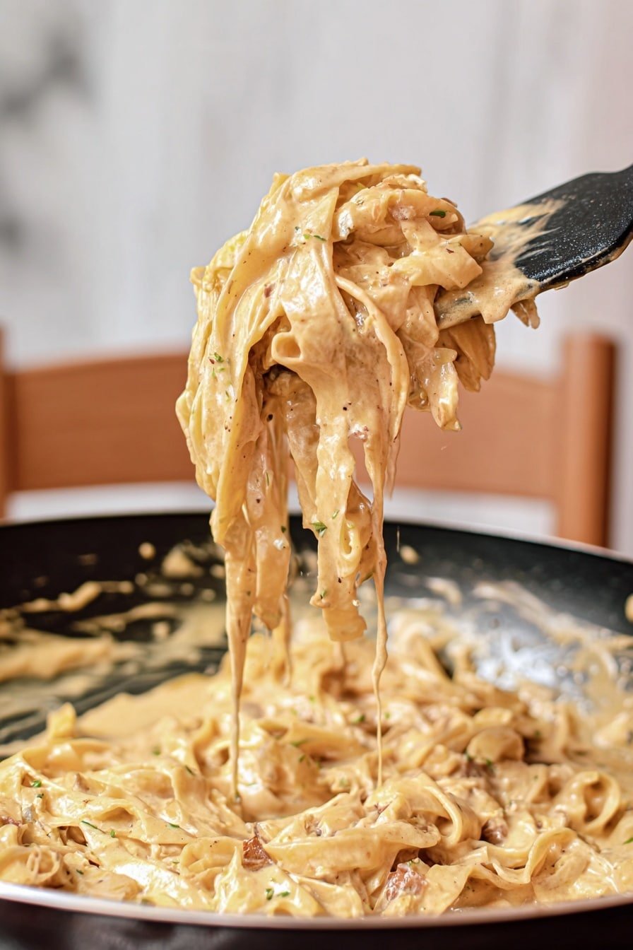 The image shows thick creamy pasta being lifted with a black spatula over a black pan filled with more pasta. The pasta is coated in a smooth, light beige creamy sauce with visible small bits of seasoning and darker pieces that look like mushrooms or herbs. The background has a soft, white marbled texture, and the scene includes a blurred wooden chair, adding a warm, homey feel. The woman's hand holding the spatula is partly visible at the right side of the image. photo taken with an iphone --ar 2:3 --v 7