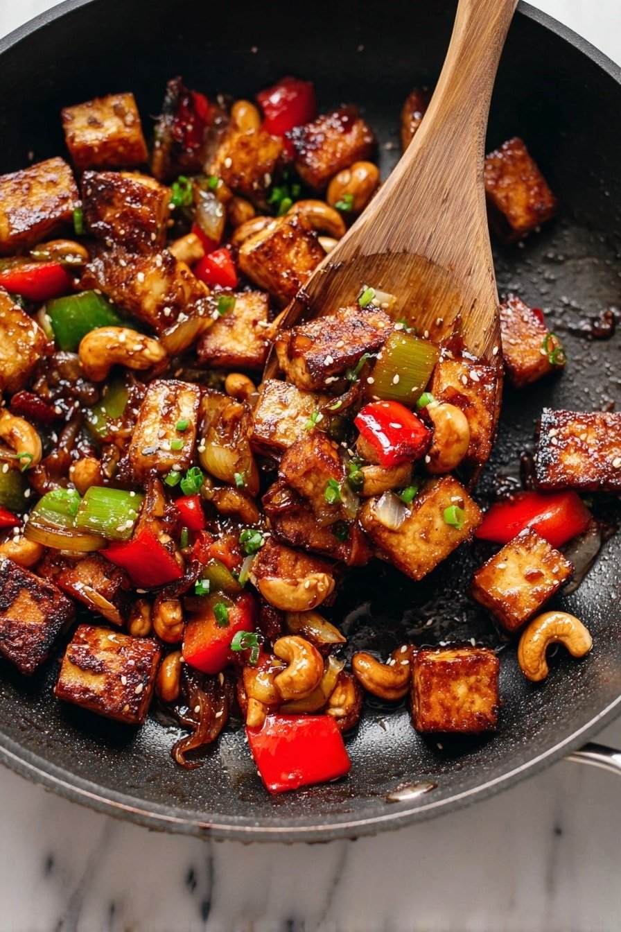 The image shows a metal pan filled with crispy golden-brown tofu cubes that are slightly charred on the edges. Mixed in with the tofu are chopped pieces of red and green bell peppers, light brown cashew nuts, and translucent cooked onion chunks. The ingredients look shiny with a dark, thick sauce. A wooden spoon is scooping up a mix of tofu, peppers, and cashews within the pan. Small green herb pieces and sesame seeds are sprinkled over the tofu, adding a fresh and textured look. The pan sits on a white marbled surface. photo taken with an iphone --ar 2:3 --v 7