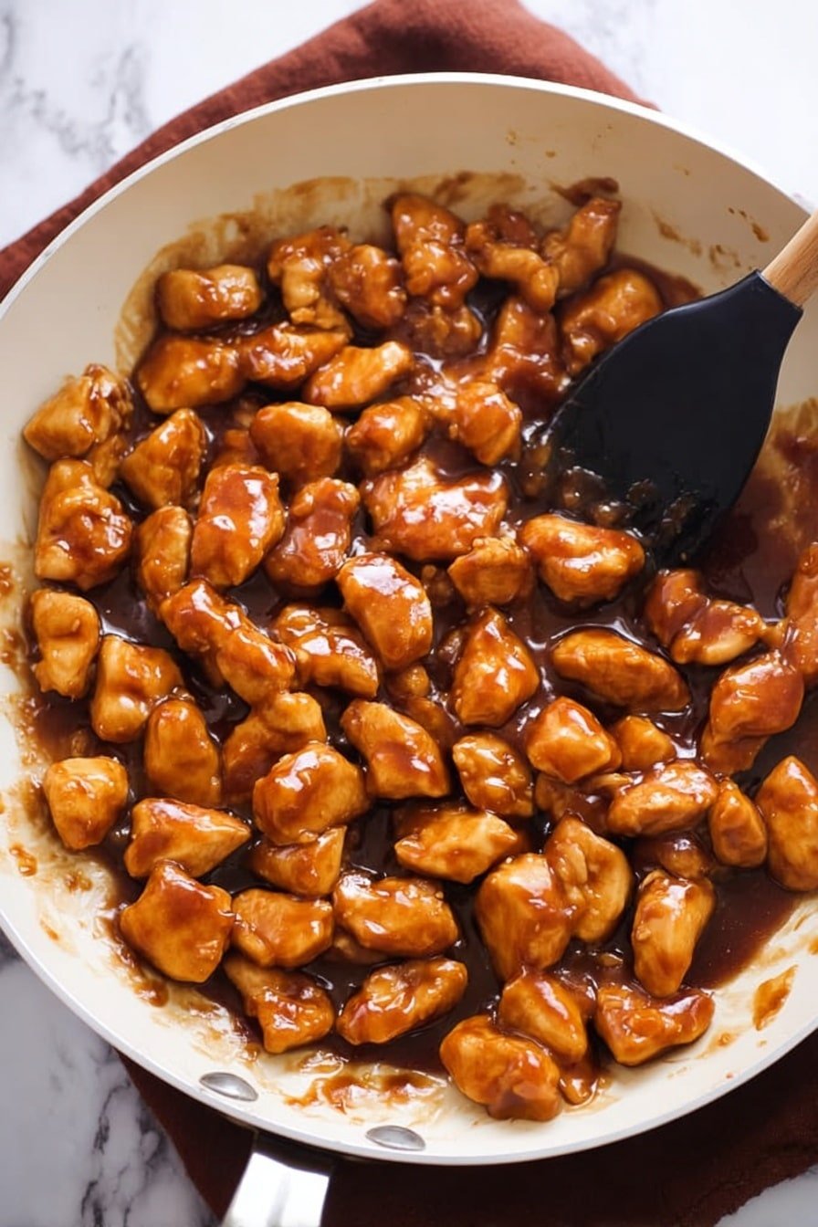 The image shows a white frying pan filled with many small, shiny pieces of golden-brown cooked meat covered in a thick, dark brown sauce. The sauce looks smooth and glossy, evenly coating the meat chunks. A wooden spatula is stirring the mixture from the right side of the pan. The pan rests on a red cloth on a white marbled surface. photo taken with an iphone --ar 2:3 --v 7