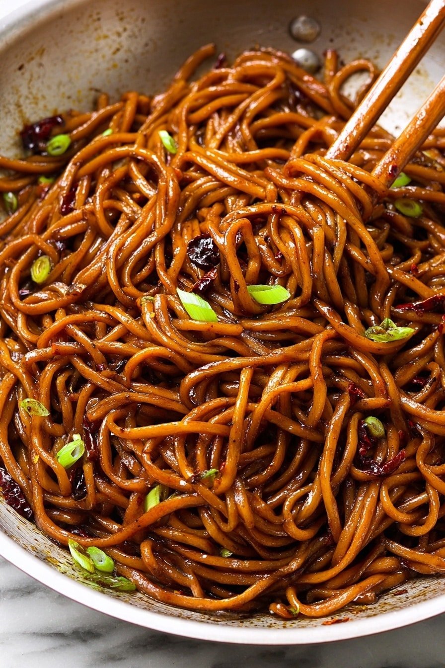 A close-up view of a bowl filled with dark brown noodles coated richly in sauce, showing a shiny, slightly oily texture. Scattered around the noodles are small pieces of green onion, adding bright green spots, and bits of dark red chili flakes spread throughout. Two wooden chopsticks are lifting a twisted bundle of noodles at the center, with some green onion pieces caught in the noodles. The bowl appears white, and the background is a white marbled texture. Photo taken with an iphone --ar 2:3 --v 7