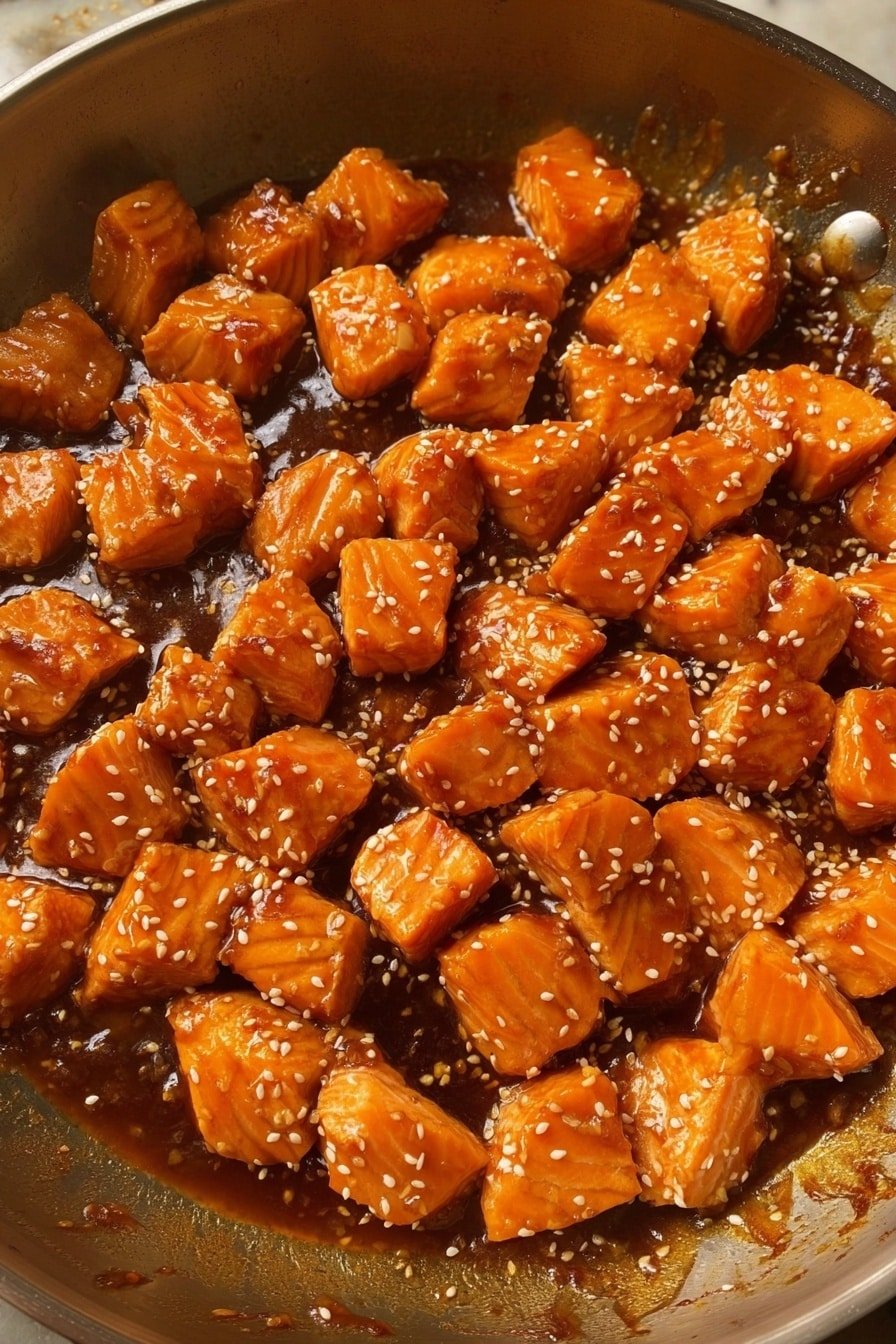 The image shows a metal pan filled with many small, golden-orange chunks of cooked salmon, all coated evenly in a thick, shiny sauce that has a rich brown color. The salmon pieces have a moist texture and are sprinkled with small white sesame seeds scattered on top. The sauce gathers at the bottom of the pan, giving a glossy and slightly sticky look. The pan itself has a slightly worn, reflective surface that contrasts with the bright food inside. photo taken with an iphone --ar 2:3 --v 7