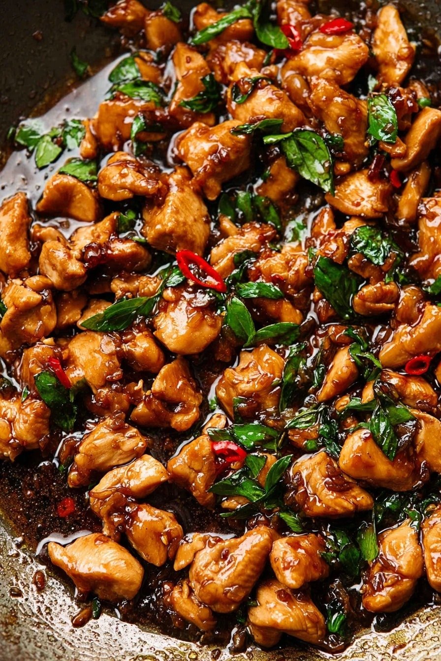 This image shows a close-up of many small pieces of cooked chicken in a dark brown sauce that looks shiny and thick, mixed with bright green basil leaves and small red chili slices. The chicken pieces are irregular in shape and size, covered evenly with the sauce which has a rich, glossy texture. The background is a dark cooking pan surface with some oil and sauce pooling around the chicken. photo taken with an iphone --ar 2:3 --v 7