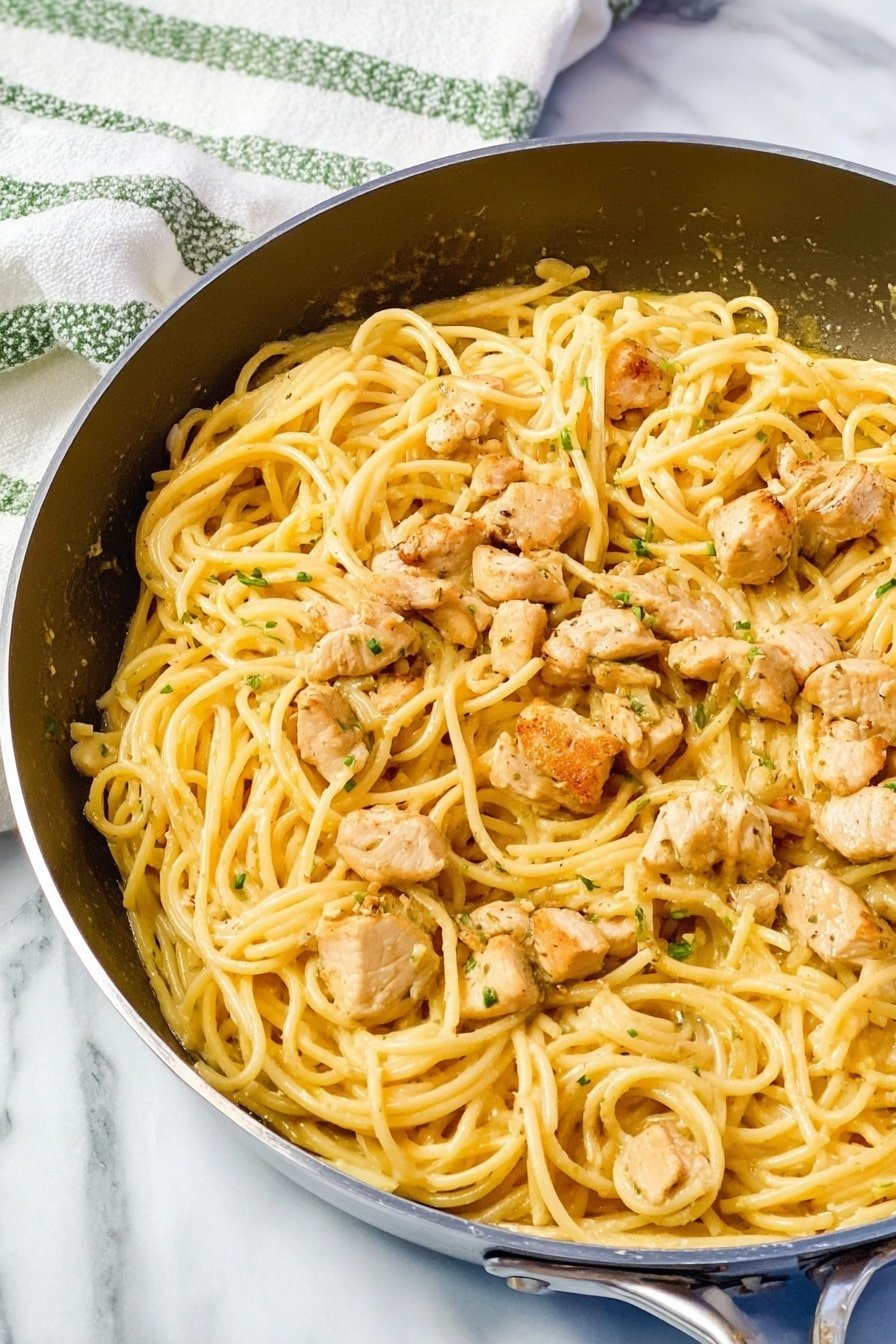 The image shows a black cast iron skillet filled with two main layers: the bottom layer is light yellow spaghetti noodles with a smooth texture, evenly spread across the pan, and the top layer consists of medium-sized, slightly browned pieces of cooked chicken, scattered over the noodles. The skillet rests on a white marbled surface, with a white and yellow striped cloth partially visible in the background. The overall colors are warm tones of beige, light yellow, and soft brown, giving a hearty and cooked look. Photo taken with an iphone --ar 2:3 --v 7