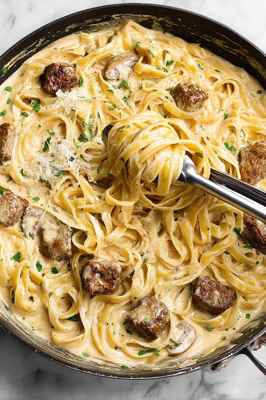 A close-up of a skillet filled with creamy fettuccine pasta mixed with browned meat chunks, all coated in a thick, pale yellow cream sauce with small green herb pieces scattered throughout. A pair of silver tongs hold a twisted portion of the pasta with a piece of meat wrapped inside. The skillet sits on a surface with a white marbled texture. photo taken with an iphone --ar 2:3 --v 7