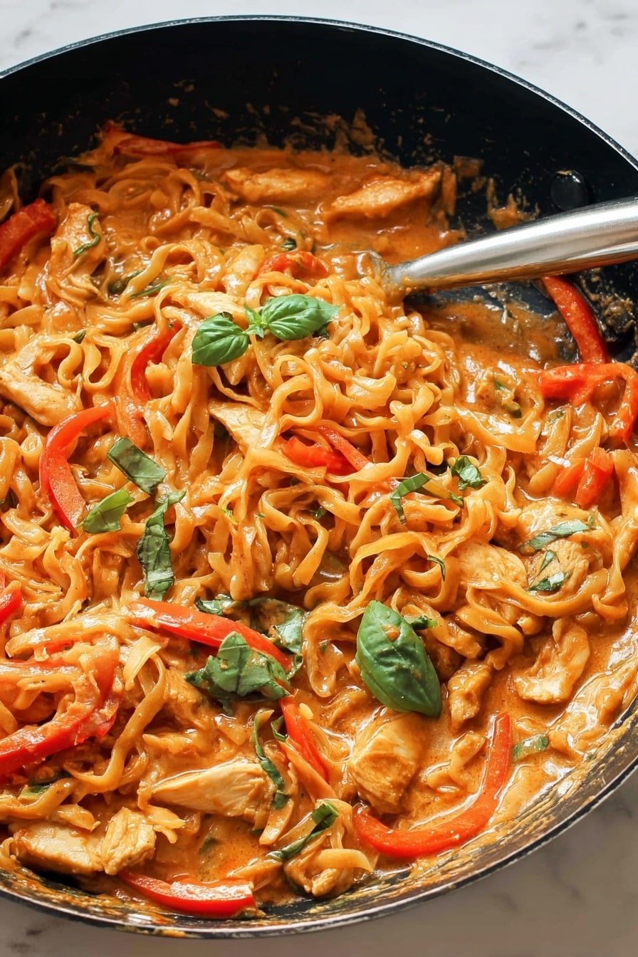 The image shows a pan filled with a rich orange sauce dish that contains wavy noodles, sliced red bell peppers, and pieces of cooked chicken. Bright green basil leaves are scattered on top, adding a fresh touch. The noodles and chicken are coated in a thick creamy sauce, and the red pepper slices stand out with their vibrant color. The pan has a black interior, and a silver cooking utensil rests inside the pan. The background features a white marbled texture. photo taken with an iphone --ar 2:3 --v 7