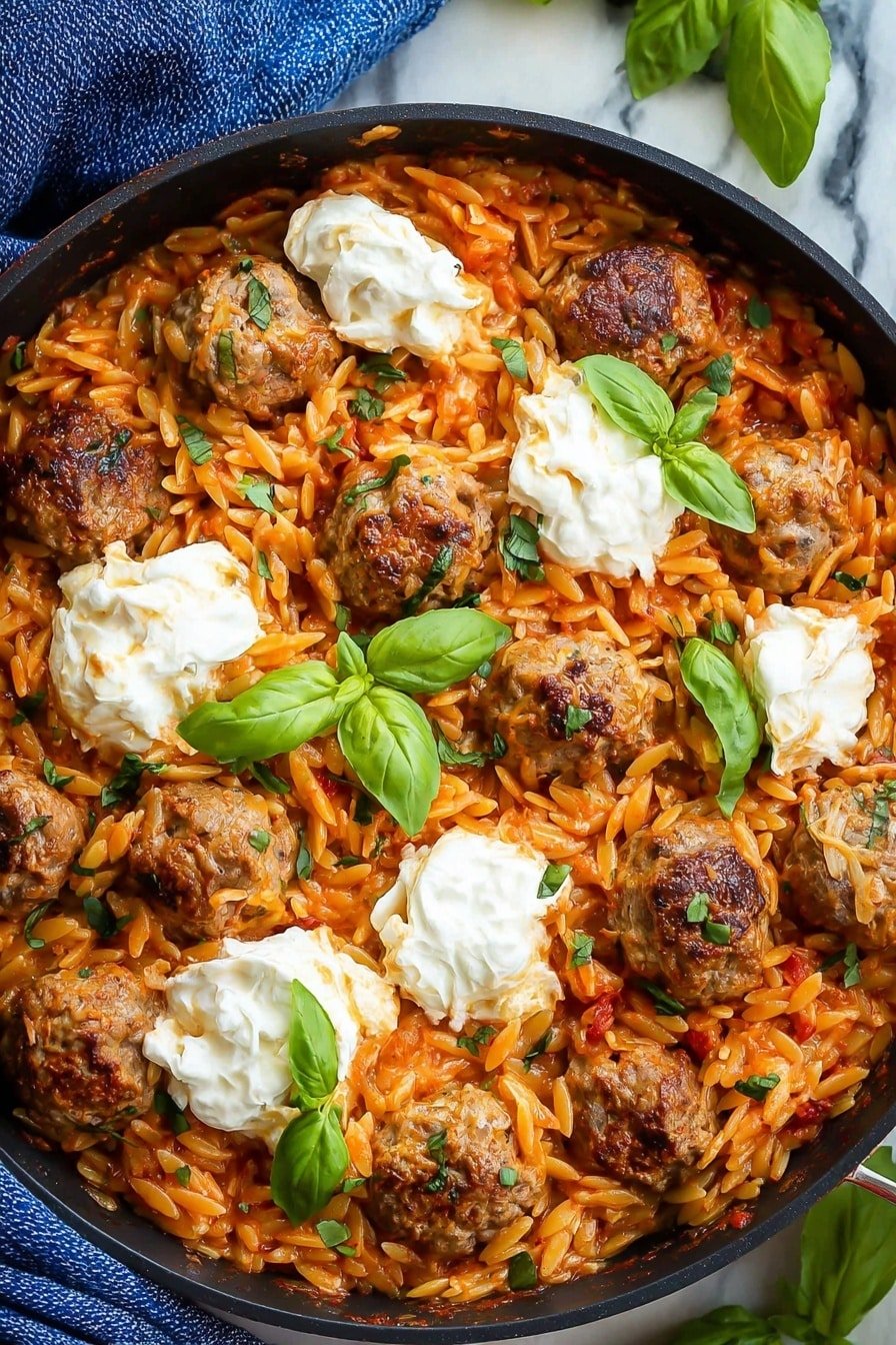 The dish is a skillet filled with a mix of orange-red orzo pasta and small browned meatballs, with visible pieces of cooked onion mixed in. On top, there are several dollops of white creamy cheese spread around, and fresh green basil leaves are placed in between the cheese and meatballs. The skillet rests on a white marbled surface with a blue cloth partially visible at the top left edge. The colors are warm and rich, with the green basil adding a fresh pop to the overall look. photo taken with an iphone --ar 2:3 --v 7
